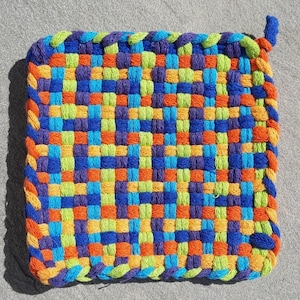 May include: A colourful woven pot holder with a square shape and a knotted handle. The pot holder is made with blue, green, orange, and yellow yarn.