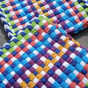 May include: Two colorful woven potholders. Each potholder is square and features a woven pattern of white, blue, purple, yellow, green, and red threads. The edges are finished with a white and blue border.