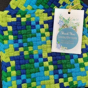May include: A woven potholder in shades of blue, green, and turquoise. The potholder has a square pattern and a loop for hanging. A tag with the text "Thank You Tuesday Afternoons" is attached.