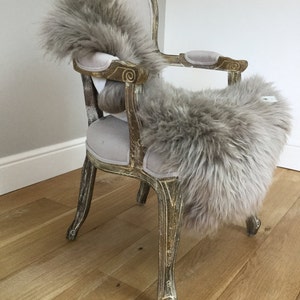 Puede incluir: Una alfombra de piel de oveja gris claro drapeada sobre una silla vintage con un asiento de terciopelo gris claro y patas ornamentadas pintadas de oro.
