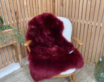 May include: A deep burgundy sheepskin rug draped over a white upholstered chair with wooden arms and legs. The chair sits on a beige woven rug, with a wooden slatted wall in the background. A small plant in a woven basket is visible.