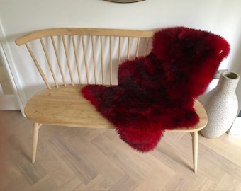 May include: A wooden bench with a red sheepskin throw draped over it. The bench has a curved back and a slatted design. The sheepskin is soft and fluffy.