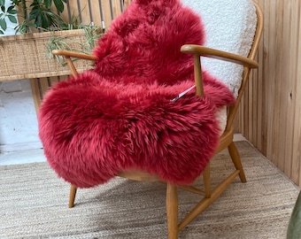 May include: A wooden armchair with a cream-colored seat cushion and a vibrant red sheepskin throw. The throw covers the seat and back of the chair, adding a touch of color and texture. The chair is set on a neutral-toned woven rug.