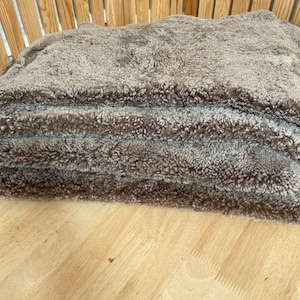 May include: A stack of three brown sheepskin rugs with a fluffy texture. The rugs are folded and placed on a wooden surface.