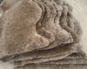 May include: A stack of brown sheepskin rugs with a wavy edge. The rugs are folded and arranged on a natural fiber rug.