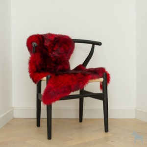 May include: A black wooden chair with a woven seat and a vibrant red sheepskin rug draped over it. The rug has a mix of red and dark brown tones, adding a touch of warmth and texture to the scene. The chair is set against a white wall.