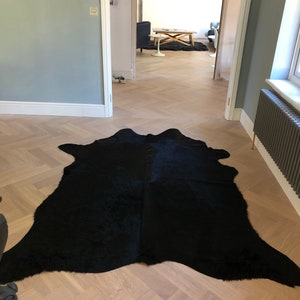 May include: A black cowhide rug on a light wood herringbone floor. The rug is in the foreground of the image, with a doorway leading to another room in the background.
