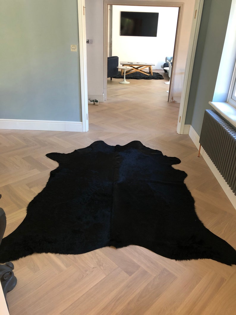 May include: A black cowhide rug on a light wood herringbone floor. The rug is in the foreground of the image, with a doorway leading to another room in the background.