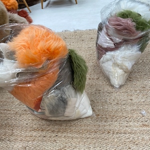 May include: Two clear plastic bags filled with various colors of faux fur. The bags are sitting on a woven rug.