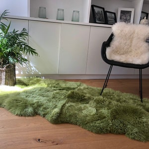 May include: A green sheepskin rug with a fluffy texture, placed on a wooden floor in a living room setting. A black chair with a white sheepskin cushion is in the background.