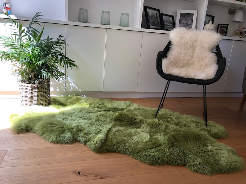 May include: A green sheepskin rug with a fluffy texture, placed on a wooden floor in a living room setting. A black chair with a white sheepskin cushion is in the background.