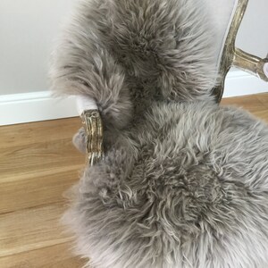 Puede incluir: Una alfombra de piel de oveja gris colocada en una silla con un asiento de tela gris claro y patas de madera tallada pintadas de oro.