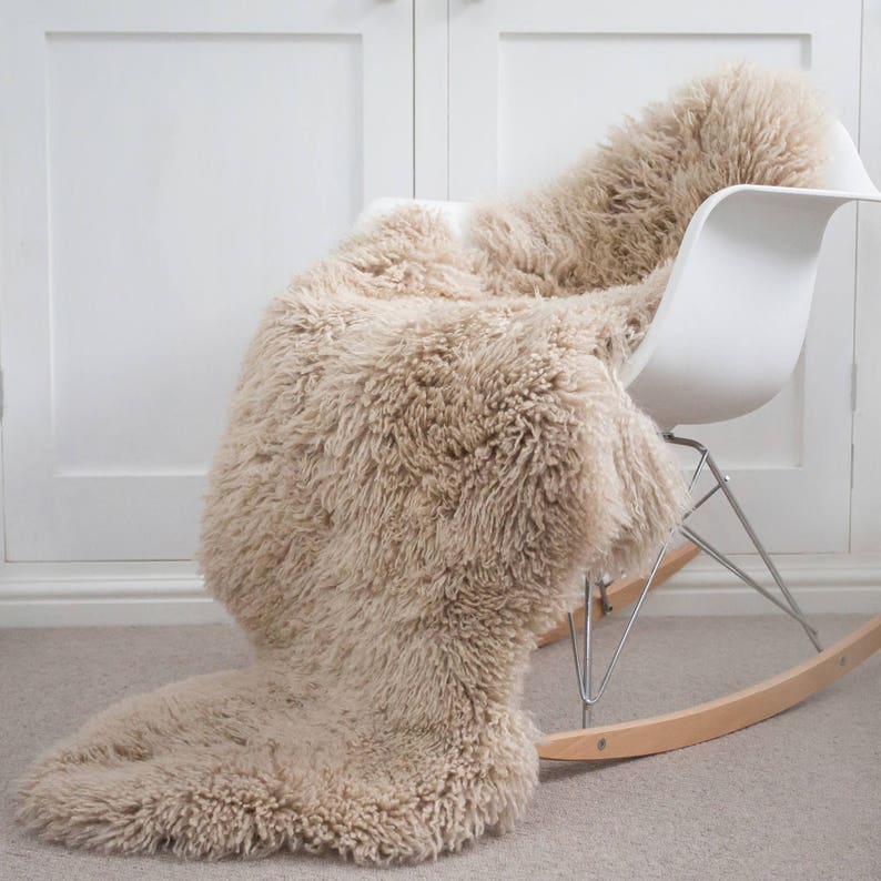 May include: A beige sheepskin rug draped over a white rocking chair. The rug has a thick, shaggy texture and a natural, neutral color. The chair has a modern design with a white seat, chrome legs, and wooden rockers.