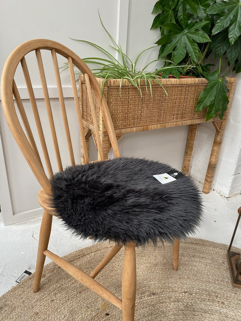 Op de afbeelding: Een houten stoel met een donkergrijs nep bont kussen. De stoel staat voor een rieten plantenstandaard met een groene plant.