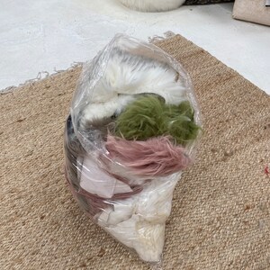 May include: A clear plastic bag filled with various colors of faux fur. The fur is white, pink, green, and brown. The bag is sitting on a woven rug.