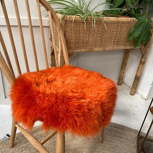 Op de afbeelding: Een feloranje schapenvacht stoelzitting. De zitting is rond en pluche, en zit op een houten stoel. De stoel staat voor een rieten plantenstandaard.