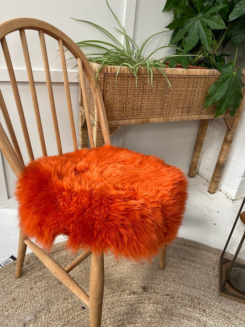 Op de afbeelding: Een feloranje schapenvacht stoelzitting. De zitting is rond en pluche, en zit op een houten stoel. De stoel staat voor een rieten plantenstandaard.