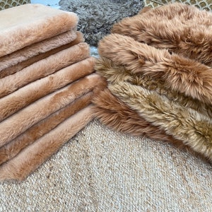 May include: A stack of light brown faux fur rugs, with a second stack of rugs in a darker brown shade.