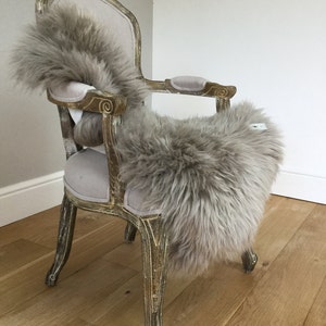 Puede incluir: Una alfombra de piel de oveja gris claro drapeada sobre una silla de estilo vintage con un asiento de tela gris claro y patas de madera talladas. La silla est&aacute; en una habitaci&oacute;n con suelo de madera clara.