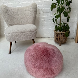 May include: A round, fluffy, light pink ottoman on a white floor. A light grey, upholstered armchair with wooden legs is in the background. A potted plant with green leaves is to the right.