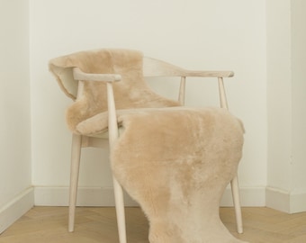 May include: A light beige sheepskin rug draped over a wooden chair. The rug is soft and fluffy, and the chair is made of light wood.
