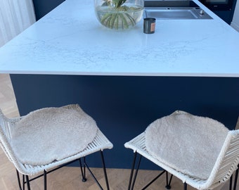 May include: Two white wicker bar stools with black metal legs and white sheepskin seat cushions. The stools are in a kitchen with a white countertop and a vase of flowers.