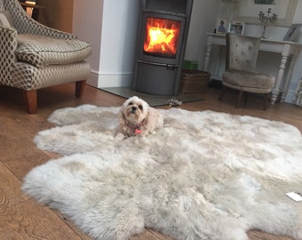 May include: A small, white dog with a pink collar is lying on a large, fluffy, white sheepskin rug in a living room. The rug is in front of a fireplace with a fire burning inside. There is a chair to the left of the rug and a table with a chair to the right.