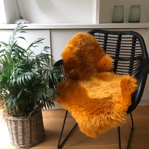 Puede incluir: Una alfombra de piel de oveja de color amarillo mostaza colocada en una silla de mimbre negra con un marco de metal negro. La silla está en una habitación con una estantería blanca y una planta en maceta.
