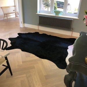 May include: A black cowhide rug on a light wood herringbone floor. The rug is in the center of the image and is surrounded by a white wall and a black radiator.