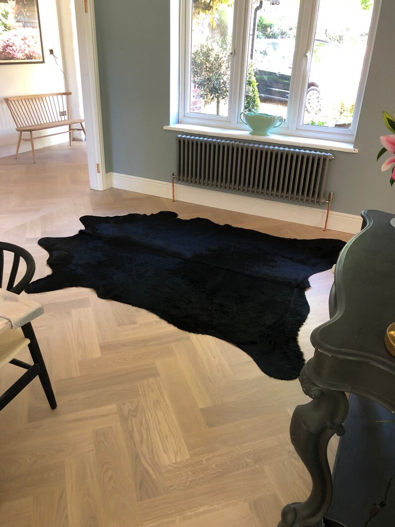 May include: A black cowhide rug on a light wood herringbone floor. The rug is in the center of the image and is surrounded by a white wall and a black radiator.