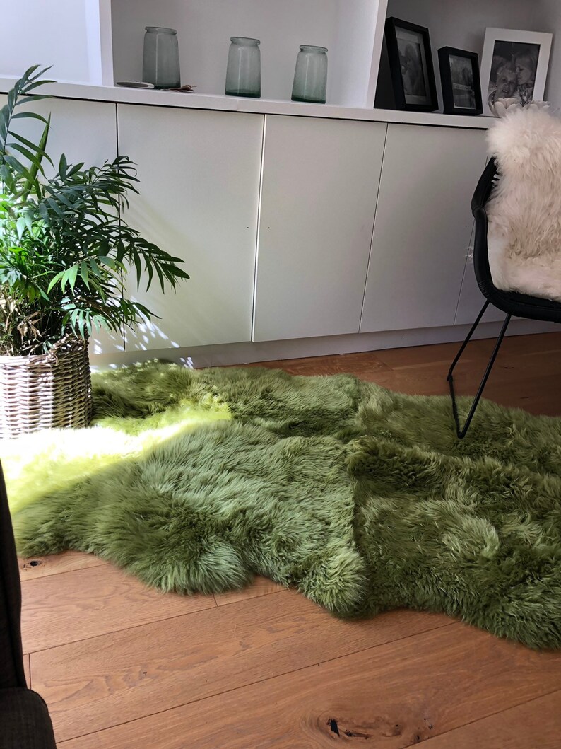 May include: A soft, green sheepskin rug with a light green gradient. The rug is laid out on a wooden floor in a living room setting.