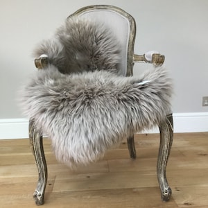 Puede incluir: Una alfombra de piel de oveja gris claro drapeada sobre una silla de estilo vintage con un coj&iacute;n de tela gris claro. La silla tiene un acabado blanco desgastado y patas ornamentadas.