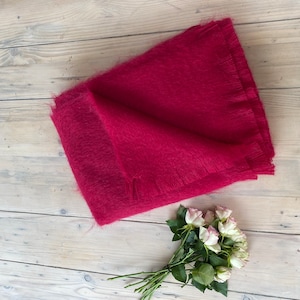 Op de afbeelding: Een gevouwen, magenta sjaal met een gefranjerde rand. De sjaal is gemaakt van een zacht, pluizig materiaal. Een klein boeket witte en roze rozen ligt op een lichtgekleurd houten oppervlak.