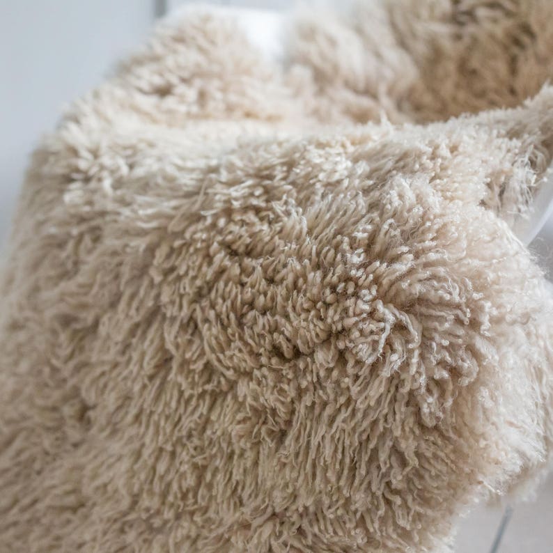 May include: Close-up of a beige, fluffy, textured blanket. The blanket has a shaggy appearance with long, soft fibers. The neutral color and plush texture suggest a cozy and comfortable home decor item.
