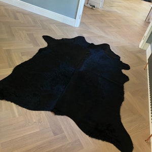 May include: A black cowhide rug with a long, flowing shape. The rug is laid out on a light wood floor with a herringbone pattern.