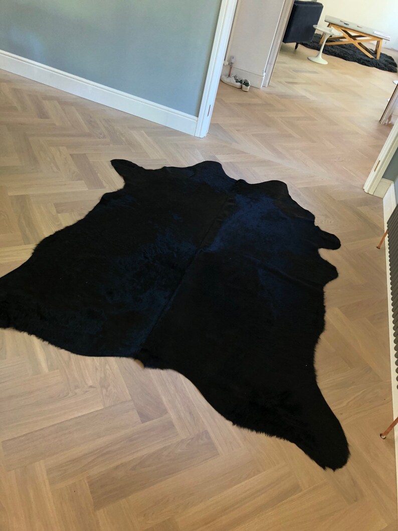 May include: A black cowhide rug with a long, flowing shape. The rug is laid out on a light wood floor with a herringbone pattern.