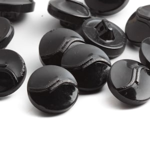 Puede incluir: Un conjunto de botones negros brillantes, ligeramente abombados. Cada botón presenta un diseño detallado en la parte delantera y un ojal en la parte trasera para coser. Los botones están dispersos sobre una superficie blanca, mostrando su calidad reflectante.