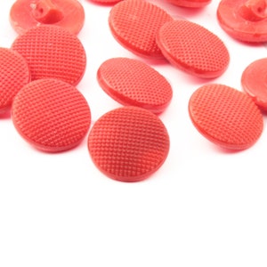 May include: A selection of red buttons in different shapes and sizes. Some are round with a textured surface, others are oval. The buttons are made of a solid, opaque material and are scattered on a white background.