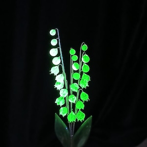May include: A luminous green glass lily of the valley sculpture against a black backdrop. The sculpture features delicate bell-shaped flowers and leaves, with a bright, glowing quality. The stems and leaves are a darker green.