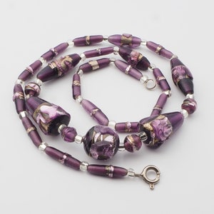 May include: A handcrafted necklace featuring a variety of purple glass beads. The necklace includes elongated, barrel-shaped, and round beads with silver accents. Some beads have gold and pink floral designs. The necklace is secured with a silver clasp.