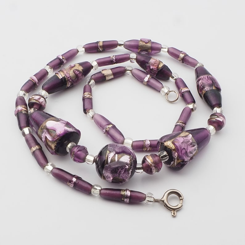 May include: A handcrafted necklace featuring a variety of purple glass beads. The necklace includes elongated, barrel-shaped, and round beads with silver accents. Some beads have gold and pink floral designs. The necklace is secured with a silver clasp.