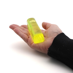 May include: A neon yellow, rectangular, translucent object is held in an open palm against a white background. The object appears to be made of a solid, smooth material. The person's arm is covered by a black sleeve.