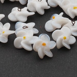Könnte beinhalten: Eine Sammlung von weißen Glasblumenanhängern mit gelben Zentren. Die Anhänger haben einen zarten, Vintage-Look und sind perfekt, um Schmuck oder Bastelarbeiten eine Prise Verspieltheit zu verleihen.