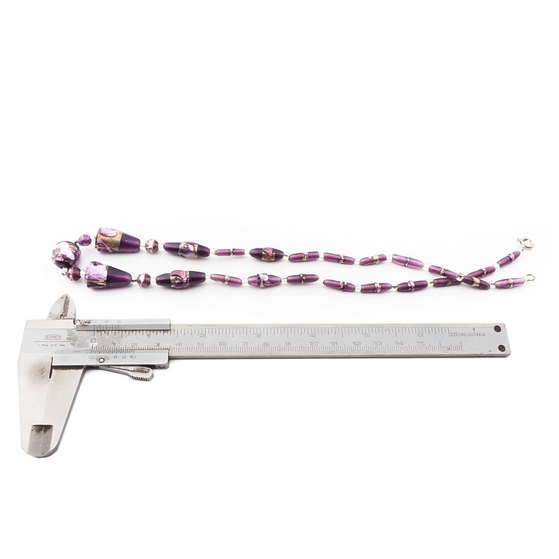 May include: A necklace with purple and gold beads of varying sizes. The necklace is laid out on a white surface with a metal caliper. The beads are a mix of round and oblong shapes, with a metallic sheen. The caliper is used to measure the necklace.