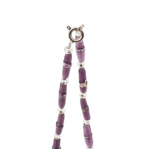 May include: A close-up of a necklace with purple beads and silver accents. The necklace features elongated, cylindrical purple beads interspersed with smaller silver beads. The clasp is silver. The necklace is on a white background.