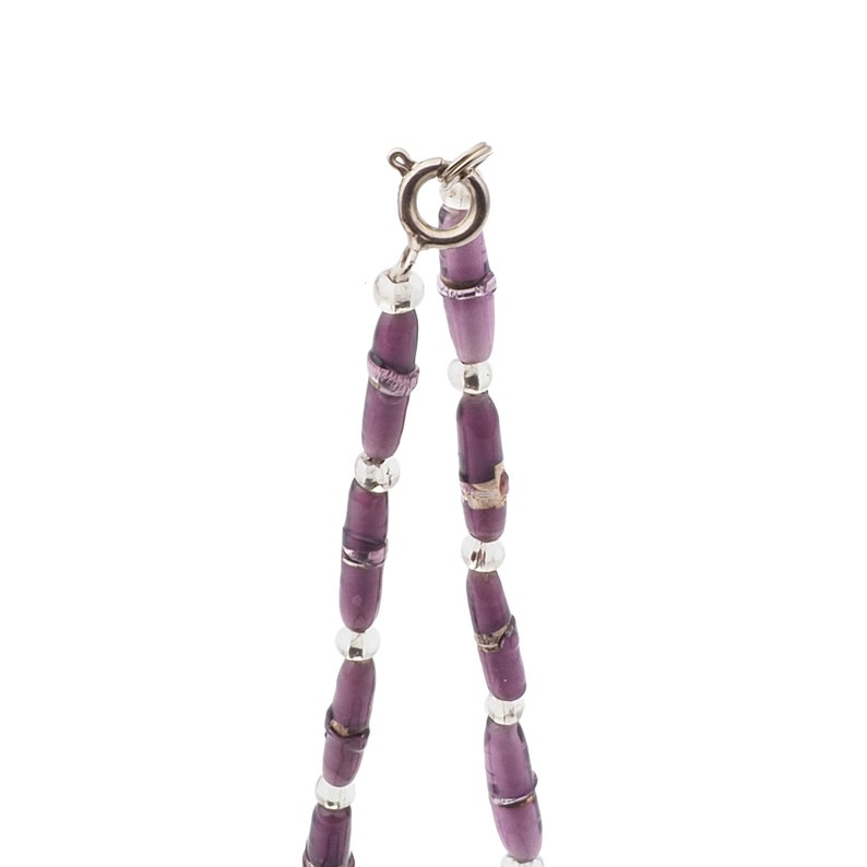 May include: A close-up of a necklace with purple beads and silver accents. The necklace features elongated, cylindrical purple beads interspersed with smaller silver beads. The clasp is silver. The necklace is on a white background.