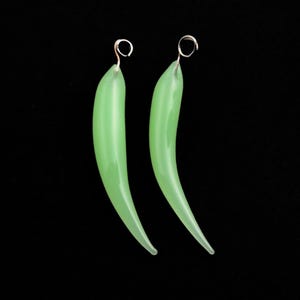 May include: A pair of green glass earrings shaped like chili peppers. The earrings have a silver loop at the top for hanging.