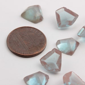 Puede incluir: Primer plano de varias cuentas de piedras preciosas facetadas e iridiscentes en tonos azules y morados. Una moneda de un centavo de cobre está al lado de las cuentas para comparar el tamaño. Las cuentas tienen forma geométrica.