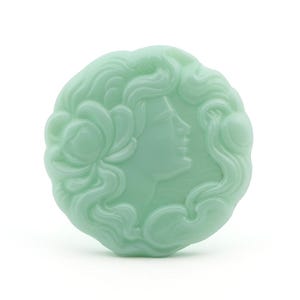 May include: A round, green glass cameo featuring a woman's profile with flowing hair and a flower design.
