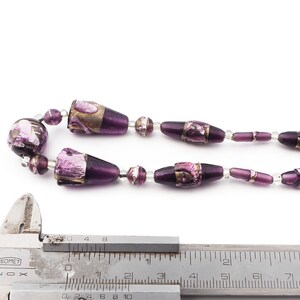 May include: A necklace featuring a series of purple glass beads with metallic accents. The beads vary in shape, including round and elongated designs, strung together with clear beads. A silver measuring tool is in the foreground.